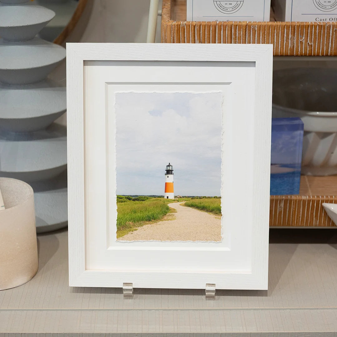 White-framed ‘Standout’ photo by Kristen Swain, styled on a shelf with pottery; lighthouse beyond a sandy path, green grass, cloudy sky.
