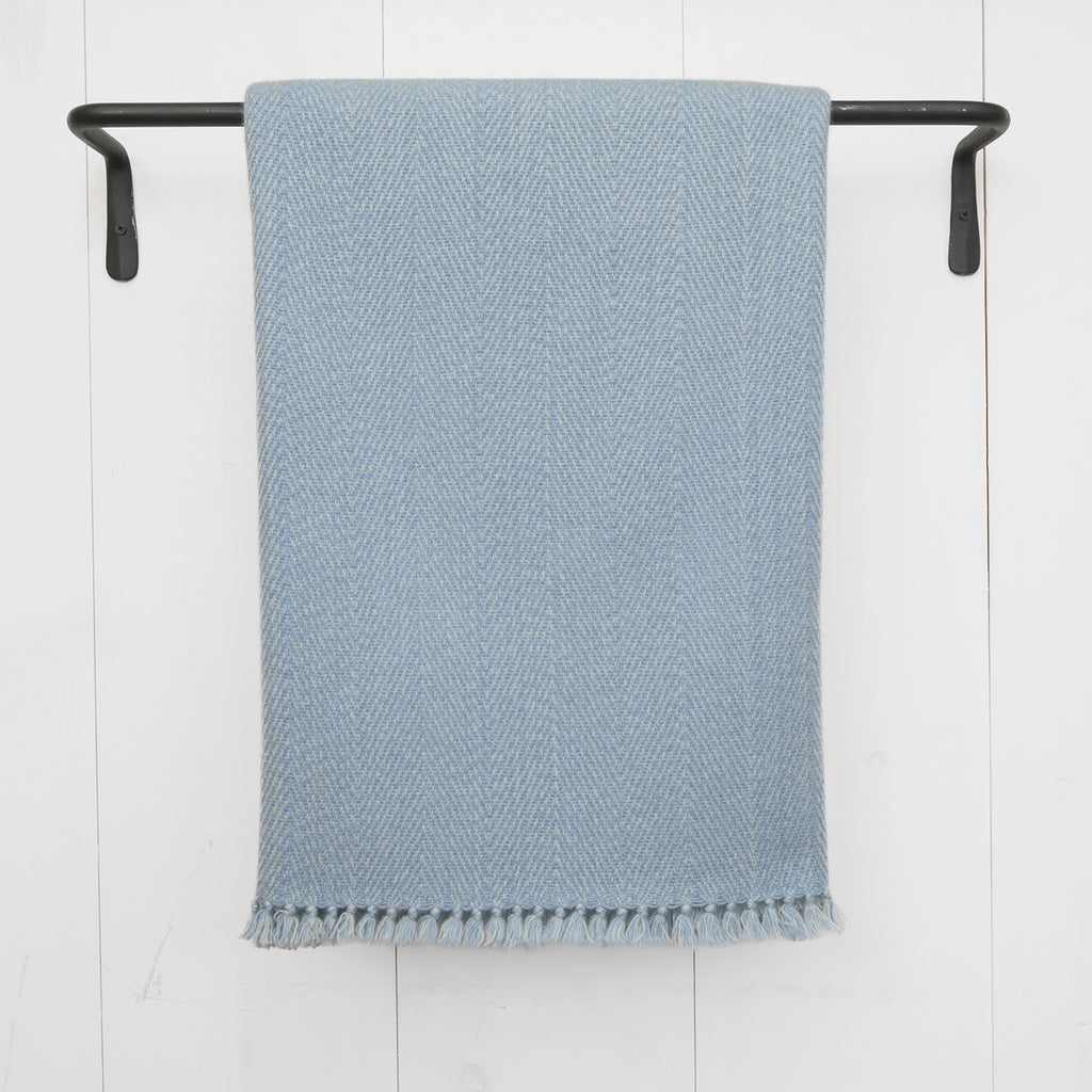 A light blue Nantucket Looms cashmere throw displayed on a black hanging rod, highlighting the delicate weave and soft fringe detailing.