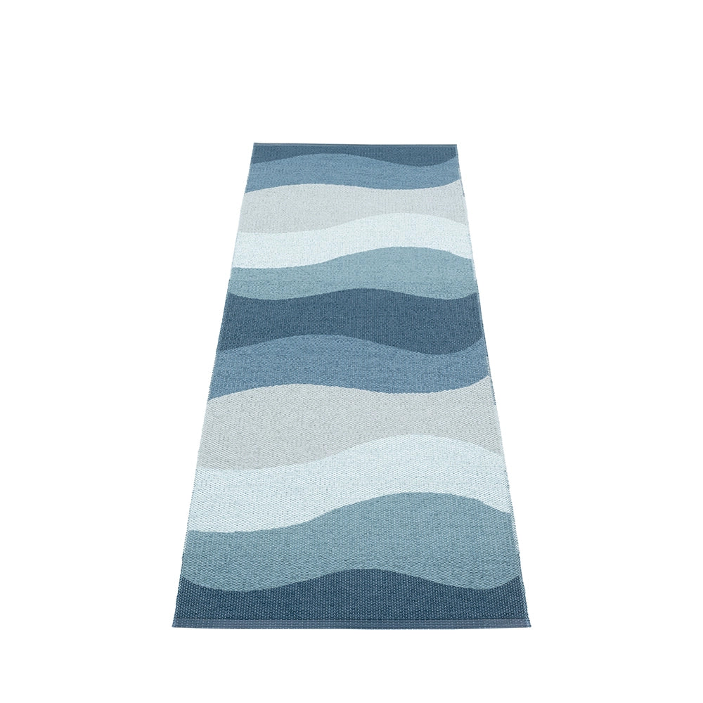 A long Pappelina rug with wavy stripes in varying shades of blue and grey, reminiscent of the ocean, laid out on a plain background.