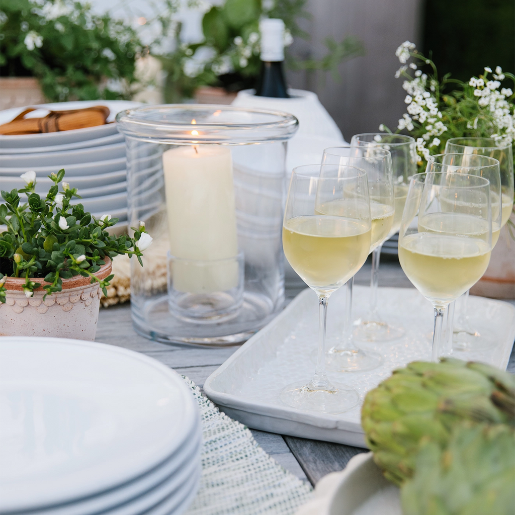 A beautifully arranged outdoor table setting featuring Nantucket Looms products. The scene includes a lit candle in a clear glass hurricane, glasses of white wine on a white tray, stacked white plates, and potted plants adding a touch of greenery. The overall ambiance is elegant and inviting, perfect for an alfresco dining experience.