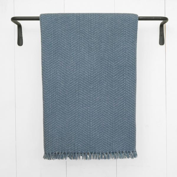 Nantucket Looms' ash blue handwoven cashmere throw draped over a black rod against a white wall, showcasing the detailed herringbone weave and fringe.