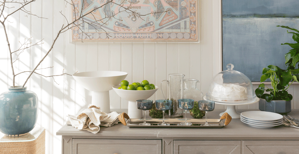 Bright and airy Nantucket dining room sideboard styled with modern glassware, a large white bowl of limes, and decorative pieces for a spring collection showcase.
